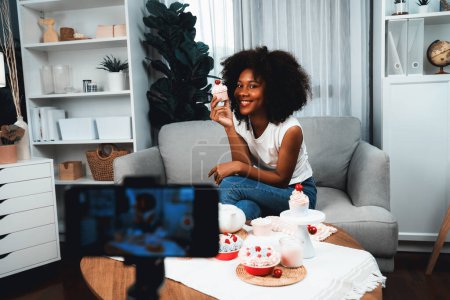 Photo for Beautiful young African blogger presenting piece of cupcake in concept special cuisine with video record on smartphone. Content creating of social media with favorite sweets bakery dish. Tastemaker. - Royalty Free Image