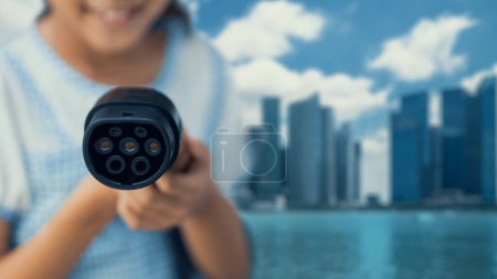 Photo for Focus EV charger pointing in front of camera with young little girls hand in blurry background with cityscape. Electric car charger plug using alternative clean energy reducing CO2 emission. Peruse - Royalty Free Image