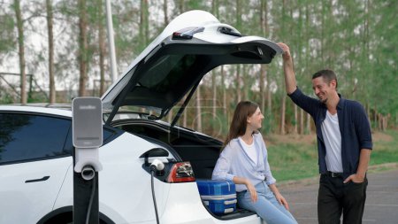 Téléchargez les photos : Couple recharger leur voiture EV avec une station de recharge de batterie électrique à l'arrêt pendant leur voyage sur la route pendant la saison automnale au parc national. Vacances relaxantes avec voiture écologique. Exalt. - en image libre de droit