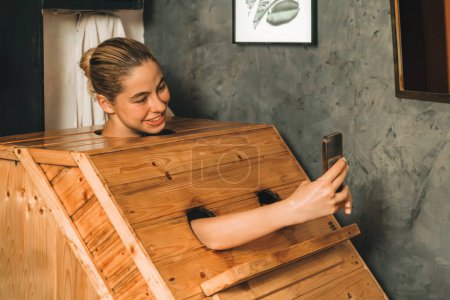 Photo for A portrait of gorgeous caucasian woman playing her mobile phone while using wooden sauna cabinet in warm tone. Attractive female with beautiful skin taking a photo. Gray background. Tranquility - Royalty Free Image