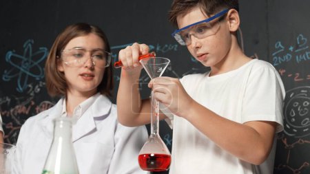 Photo for Close up of caucasian boy mixing chemical liquid while teacher giving advice. Instructor wearing lab suit looking for diverse student at table with beaker filled with colored solution. Erudition. - Royalty Free Image