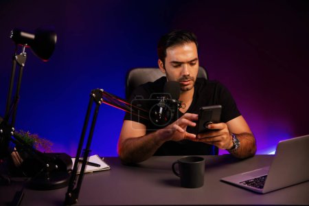 Photo for Broadcaster talking to listener on live streaming online, checking message from smartphone on social media in host channel by creative positive thinking concept at purple neon light studio. Surmise. - Royalty Free Image