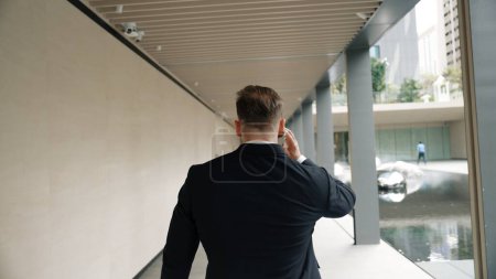 Photo for Back view of happy manager talking to phone while walking at modern corridor. Smart boss using telephone by calling marketing team to plan about financial strategy while walking at green city. Urbane. - Royalty Free Image