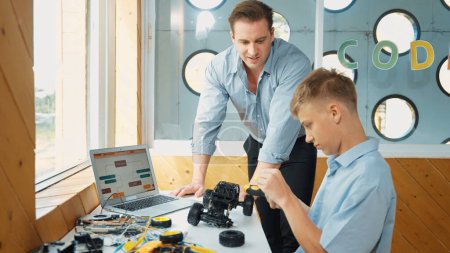 Photo for Caucasian teacher help student fixing car model and giving advise for programing system or coding prompt while highschool boy listen and use equipment to repair robot at STEM classroom. Edification. - Royalty Free Image