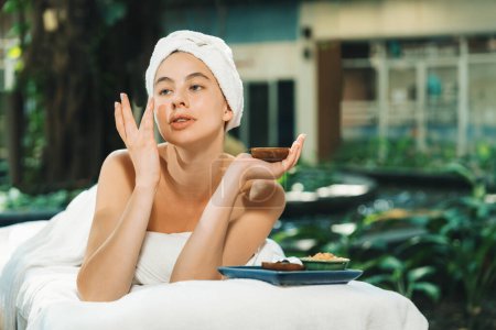 Photo for Beautiful caucasian women in white towel smells herbal homemade facial mask while lie on spa bed surrounded by outdoor natural environment. Relaxing and chilling concept. Tranquility. - Royalty Free Image