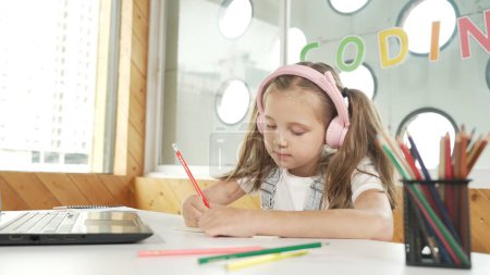 Photo for Cute smart girl doing classwork or online learning while listen to teacher. Happy student writing, drawing, working on paper while wearing headphone with laptop and colorful color pencils. Erudition. - Royalty Free Image