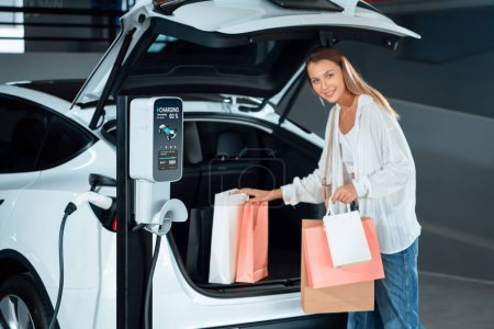 Junge Frau fährt mit Elektroauto zum Parkplatzladen eines Einkaufszentrums in der Innenstadt und zeigt urbanen nachhaltigen Lebensstil durch grüne, saubere wiederaufladbare Energie von Elektrofahrzeugen