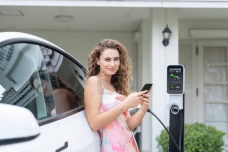Téléchargez les photos : Femme écologique moderne rechargeant le véhicule électrique de la station de charge EV à la maison. Utilisation innovante de la technologie EV pour suivre l'utilisation de l'énergie afin d'optimiser la charge de la batterie à la maison. Synchronos - en image libre de droit