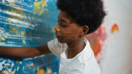 Photo for Side view of happy african boy painted the stained wall with colorful hand while wearing casual white shirt in art lesson.Smart student use hand print to make creative artwork. Education. Edification. - Royalty Free Image
