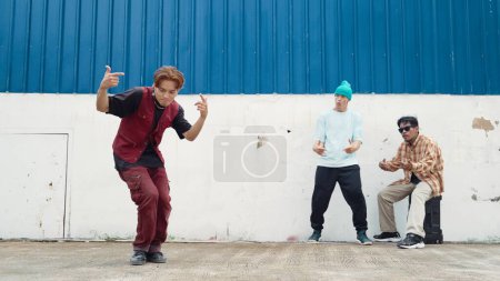 Photo for Young street dancer dancing in hip hop style with multicultural friends cheering behind at wall. Attractive handsome man stretch arms at street with blue background. Outdoor sport 2024. Endeavor. - Royalty Free Image