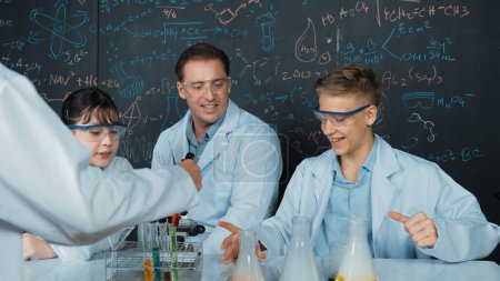 Photo for Creative teacher pointing chemistry at blackboard while talking to highschool boy at table with microscope and test tube with colored liquid. Young student wearing lab coat in STEM class. Edification. - Royalty Free Image