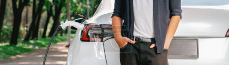 Photo for Man recharging battery for electric car during road trip travel EV car in natural forest or national park. Eco friendly travel during vacation and holiday. Panorama banner Exalt - Royalty Free Image