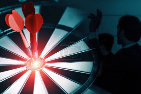 Business Target Goal For Success Strategy Concept - Red dart arrow hitting center goal on dart board with business people working in background showing precision and success of business target. uds
