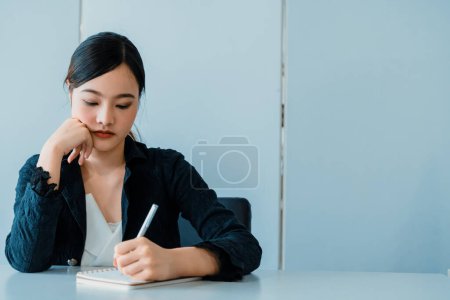 Foto de Hermosa mujer asiática joven escribe carta en el cuaderno mientras está sentado en el escritorio de la oficina. Concepto de trabajo de escritor de contenido y secretario. BARROS - Imagen libre de derechos