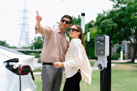 Photo for Young couple with coffee pay for electricity while recharge EV car battery at charging station connected to electrical power grid tower as electric industrial for eco friendly car travel. Expedient - Royalty Free Image