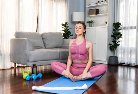 Téléchargez les photos : Femme flexible et dextérité en tenue de sport faisant position de yoga en posture de méditation sur tapis d'exercice à la maison. Mode de vie sain de yoga de gaieté à domicile avec l'esprit paisible et la sérénité. - en image libre de droit