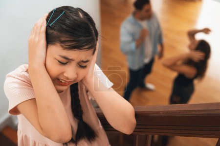 Stressed and unhappy young girl cover her ears blocking sound tension by her parent argument from the stair. Unhealthy family and domestic violence lead to traumatic childhood and anxiety. Synchronos