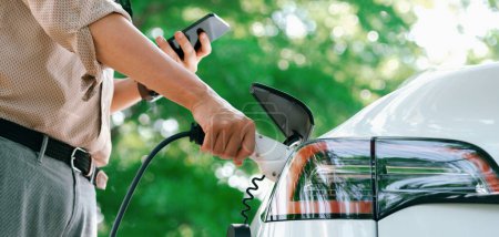 Photo for Young man use smartphone to pay for electricity at public EV car charging station green city park. Modern environmental and sustainable urban lifestyle with EV vehicle. Panorama Expedient - Royalty Free Image
