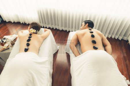 Téléchargez les photos : Massage aux pierres chaudes au salon de spa dans un complexe de luxe avec une lumière du jour sérénité ambiante, un couple heureux client profitant d'un massage aux pierres de basalte spa glisse sur le corps avec une chaleur apaisante. Tranquillité - en image libre de droit