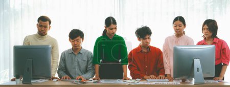 Photo for Young asian startup group works together on a project. Professional business people determine and focus on their performance while standing at office room with office equipment placed. Variegated. - Royalty Free Image