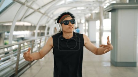 Foto de Primer plano del guapo bailarín inteligente moviendo las manos mientras baila música hip hop. Hipster asiático con auriculares mientras camina por el pasillo con un fondo borroso. Deporte al aire libre 2024. Rígidamente.. - Imagen libre de derechos