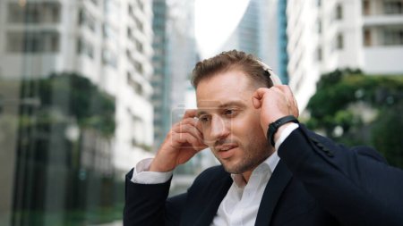 Businessman put on headphone and listen relaxing music while sitting at green city with blurred background . Skilled manager project manager enjoy to listen while dance to song in lively mood. Urbane.