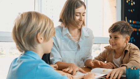 Photo for Professional caucasian teacher telling story to diverse student while sitting at table with storybook and colored book. Smart learner listening story while colored picture from instructor. Erudition. - Royalty Free Image