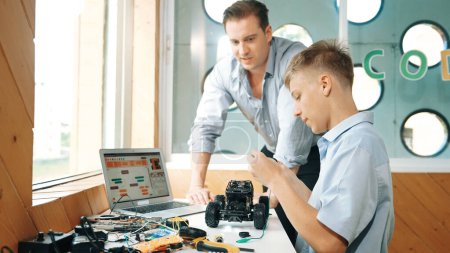 Photo for Caucasian teacher help student fixing car model and giving advise for programing system or coding prompt while highschool boy listen and use equipment to repair robot at STEM classroom. Edification. - Royalty Free Image