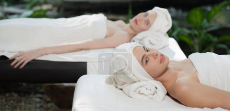 Téléchargez les photos : Deux belles jeunes femmes allongées sur un lit de spa avec serviette blanche tout en abattant dans une profonde détente arrondie par une nature relaxante et apaisante. Concept santé et beauté. Fond flou. Tranquillité. - en image libre de droit