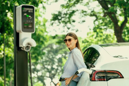 Junge Frau lädt Akku für Elektroauto während der Autoreise Elektroauto in Naturwald oder Nationalpark. Umweltfreundliches Reisen im Urlaub und im Urlaub. Erhöhen