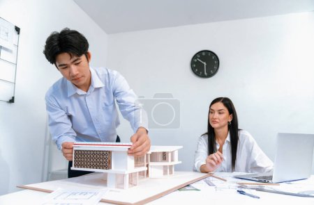 Architecte asiatique masculin professionnel utilisant la règle pour mesurer la longueur du modèle de maison tandis que jeune beau collègue caucasien utilisant un ordinateur portable pour analyser les données sur la table de réunion avec le modèle de maison. Immaculée.