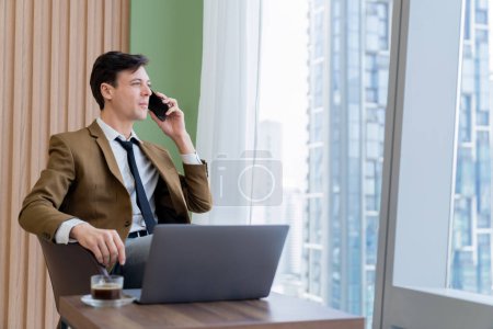 Foto de Primer plano del hombre de negocios guapo haciendo una llamada telefónica con el gerente mientras está sentado cerca de la ventana con vista al rascacielos. Gerente ejecutivo hablando de trabajo mediante el uso de teléfono y portátil. Mira a un lado. Adornado. - Imagen libre de derechos