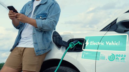 Téléchargez les photos : L'homme asiatique utilise le téléphone tout en rechargeant l'énergie de la voiture électrique, montrant l'hologramme d'état de la batterie numérique du chargeur EV de la station de charge au parc d'éoliennes. Utilisation alternative d'énergie propre. - en image libre de droit