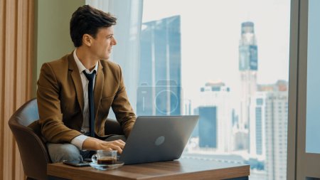 Photo for Businessman sitting on furniture working on laptop at ornamented corporate waiting area with cityscape background on the window. Business profession and strategic marketing plan for business success. - Royalty Free Image