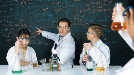 Photo for Smart teacher explain about chemical theory while student mixing colored solution or doing experiment. Instructor teaching chemistry while pointing at blackboard in STEM science class. Edification. - Royalty Free Image