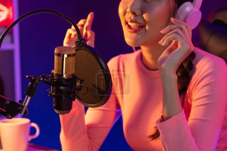 Téléchargez les photos : Chaîne hôte de jeune beau diffuseur asiatique écoutant la musique sur les médias sociaux avec les auditeurs, portant des écouteurs pastel, en utilisant un microphone pour chanter au studio intérieur néon lumière. Stratagème. - en image libre de droit