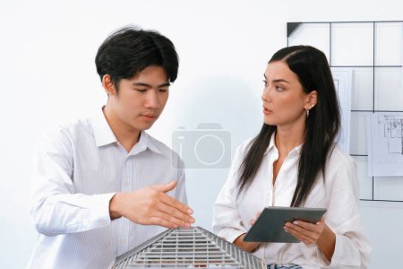 Photo for Beautiful young caucasian project manager tells asian engineer about building detail by using tablet while engineer measuring house model on meeting table with blueprint scatter around. Immaculate - Royalty Free Image