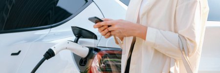 Photo for Young woman use smartphone to pay for electricity at public EV car charging station green city park. Modern environmental and sustainable urban lifestyle with EV vehicle. Panorama Expedient - Royalty Free Image