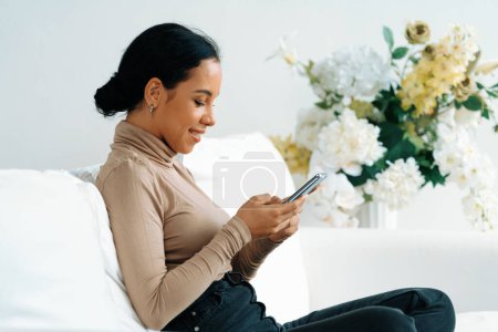 Foto de Mujer afroamericana joven y relajada usando un teléfono móvil crucial en el sofá sofá en la sala de estar en casa - Imagen libre de derechos