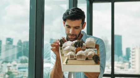 Photo for Businessman in casual outfit holding house model while checking house construction. Architect engineer inspect building model while standing near window with skyscraper. Engineering. Tracery - Royalty Free Image