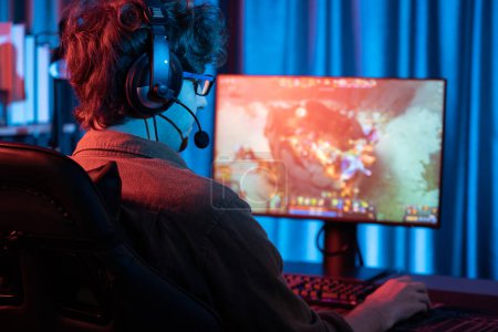 Host-Kanal von jungen Gaming-Streamer spielen Kampf Moba in der Schlacht Arena Spiel mit Multiplay-Team, trägt Kopfhörer auf dem PC-Monitor mit Rückseite Bild in Neon-Digitallicht modernen Raum. Gusher.