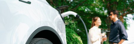 Photo for Young couple recharge electric car battery from charging station in green city park in springtime. Rechargeable EV car for sustainable environmental friendly urban travel lifestyle. Panorama Expedient - Royalty Free Image
