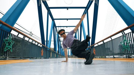 Téléchargez les photos : Hipster effectuer b-boy pas et la danse de rue au pont avec fond. Le danseur professionnel de pause porte un tissu élégant et regarde la caméra tout en posant à la caméra. Sport de plein air 2024. Brillamment. - en image libre de droit