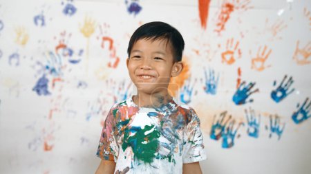 Photo for Asian happy boy looking at camera while attend in art lesson. Smart caucasian student standing in front white background while smiling at camera in lively mood. Creative activity concept. Erudition. - Royalty Free Image