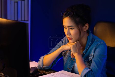 Photo for Stressful young beautiful asian creative looking on pc to search project creator social media online with planning channel strategy reach of more viewer home at neon modern office at night. Stratagem. - Royalty Free Image