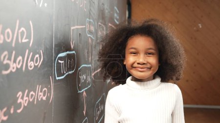 Photo for Site view of young african girl looking at camera while standing at blackboard with engineering prompt. Student smiling at camera system code written behind in STEM technology classroom. Erudition. - Royalty Free Image