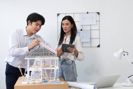 Photo for Beautiful young caucasian project manager tells asian engineer about building detail by using tablet while engineer measuring house model on meeting table with blueprint scatter around. Immaculate - Royalty Free Image
