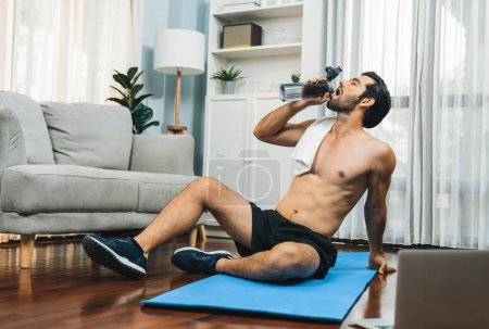 Photo for Athletic and sporty man drinking water on fitness mat after finishing home body workout exercise session for fit physique and healthy sport lifestyle at home. Gaiety home exercise workout training. - Royalty Free Image