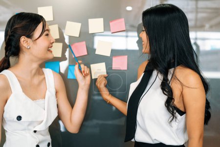Photo for Two young beautiful Asian businesswomen talk and discuss creative post on the window. It is project reminder and thinking idea notes. uds - Royalty Free Image