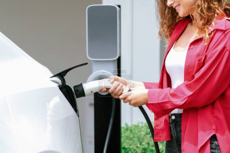 Téléchargez les photos : Femme écologique moderne rechargeant le véhicule électrique de la station de charge EV à la maison. Technologie de voiture EV utilisé pour les résidents de la maison à la durabilité environnementale future. Synchronos - en image libre de droit